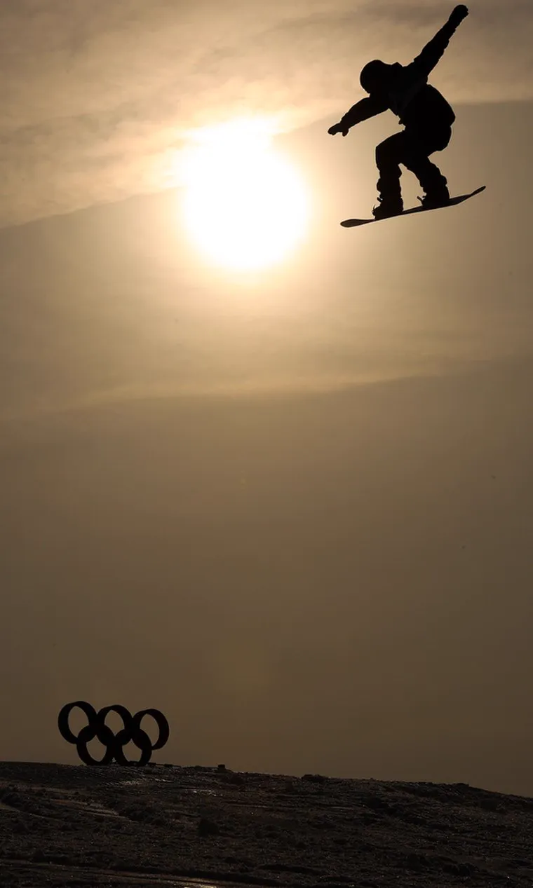 Snowboardeur en plein saut à contre-jour face au soleil, anneaux olympiques en arrière-plan